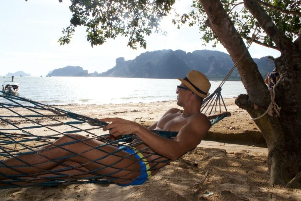 A man wearing a hat, sunglasses and blue speedo on a hammock enjoying the beautiful view of a beach and hills covered with vegetation