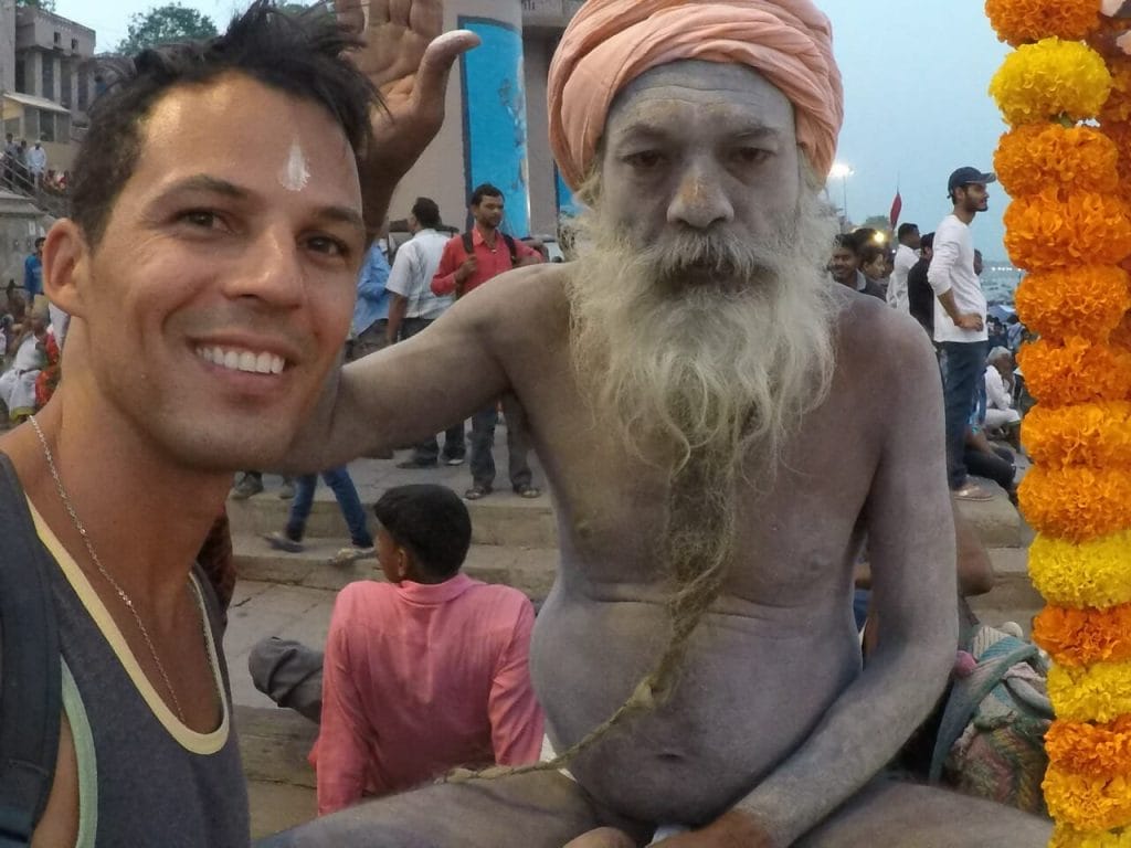 uma homem usando uma regata cinza e um colar de prata com um Sadhu que tem o corpo coberto de cinzas usando um turbante cor de salmão nas margens do rio Ganges em Varanasi, Índia