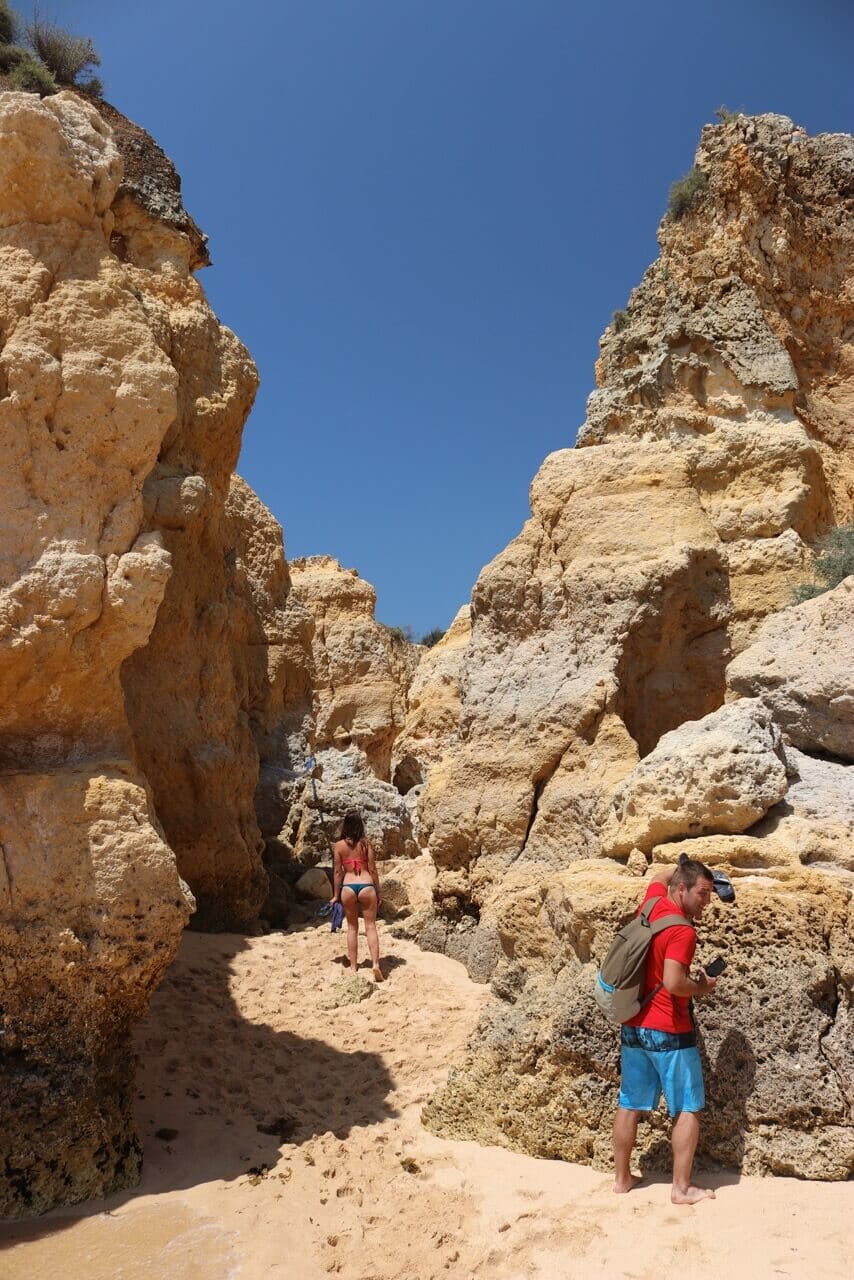 Um mulher de biquini passando pelo meio das falésias e um rapaz de camiseta vermelha, short azul e mochila próximo às falésias, Praia de São Rafael, Albufeira, Portugal