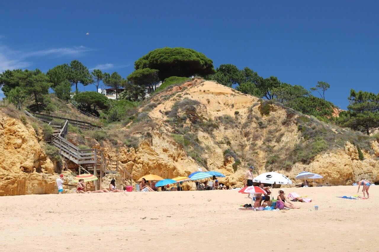 Pinheiros em cima das falésias amarelas e alguns guarda-sois e pessoas na areia, Praia de Santa Eulália, Albufeira 