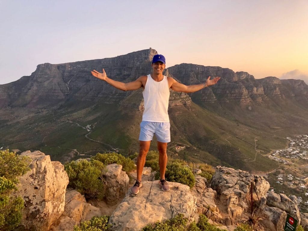 Pericles Rosa wearing a marine blue cap, white tank top, light blue shorts and sneakers at the summit of Lion's Head, Cape Town, with the Table Mountain and the Twelve Apostles in the background 