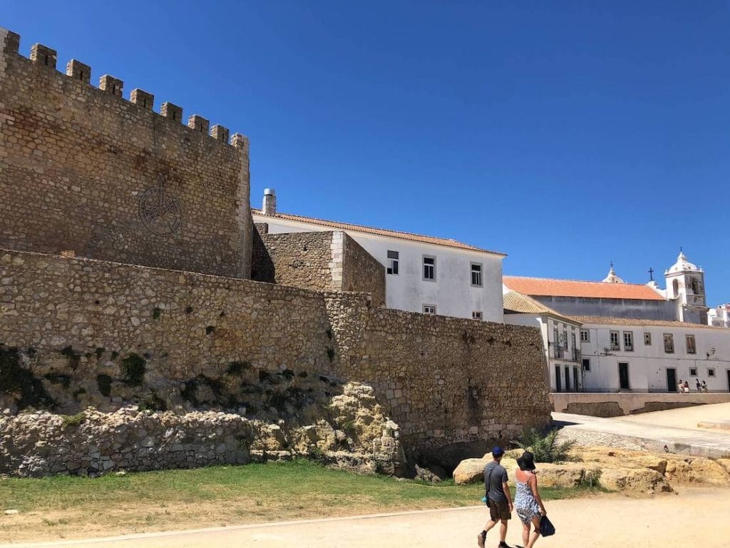 Um homem e uma mulher caminhando perto das paredes rochosas do castelo de Lagos em direção a uma igreja branca no centro da cidade de Lagos, Portugal