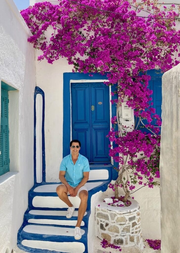 Pericles Rosa posing for a picture in front of house with adorned with a blue door and bougainvillea flowers in  Chora, Astypalea, Greece